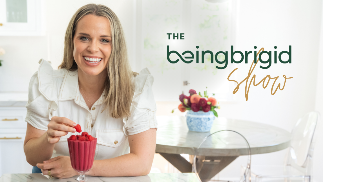 Smiling woman in a bright kitchen holding a glass filled with raspberries, seated at a table. Text on the right reads “The beingbrigid show,” with a bouquet of flowers on the table in the background.